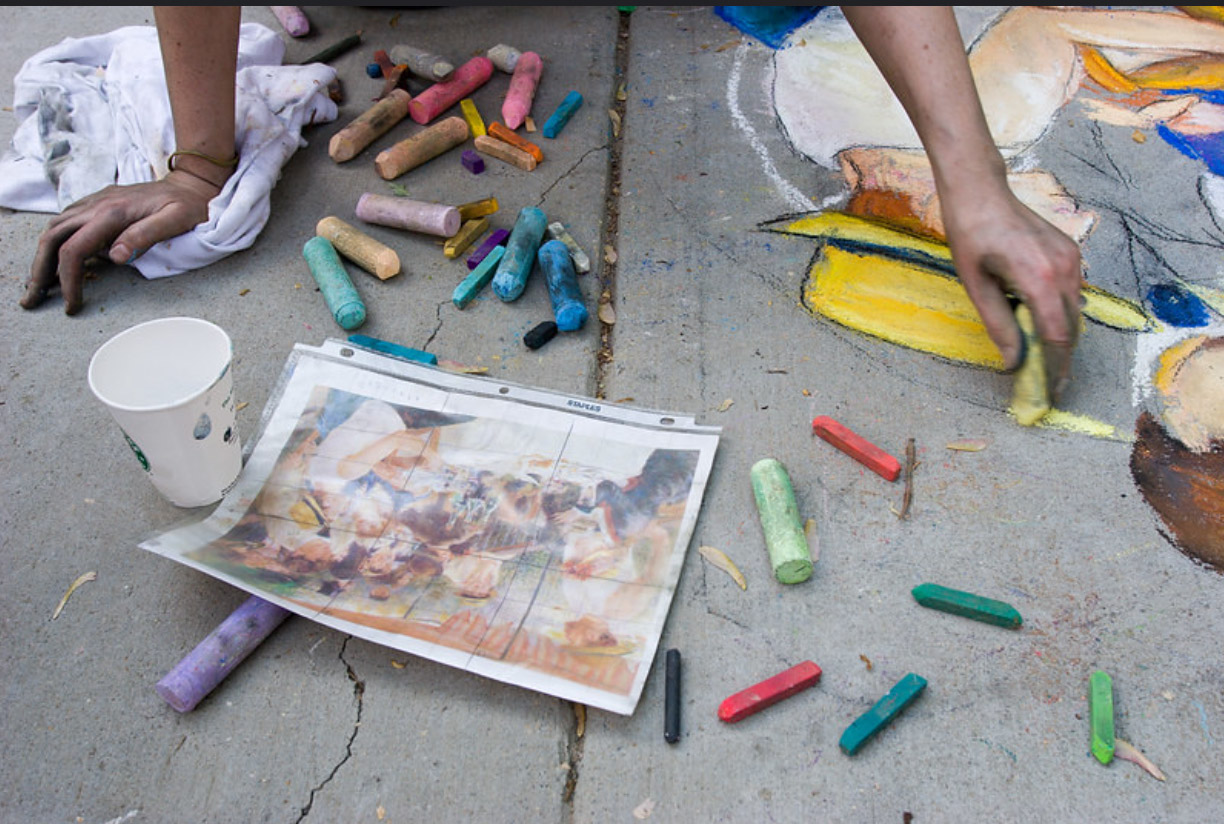 A gridded reference photo pinned to the sidewalk beside scattered pastel chalk, guiding Dana Ellyn's recreation of the Renoir