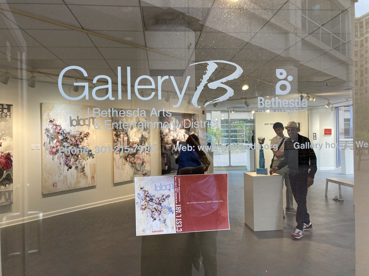 The Gallery B window in Bethesda, Maryland, with the c'est l'art exhibition poster displayed and artwork visible inside