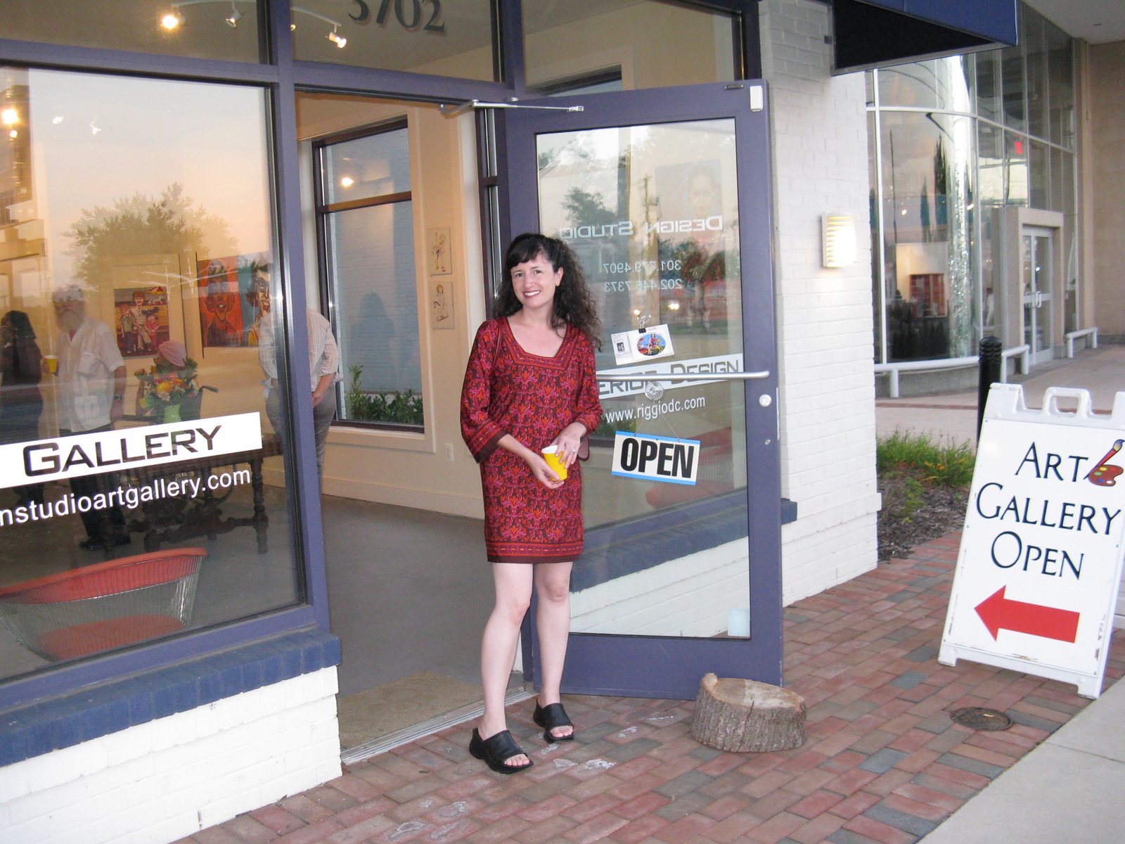 Dana Ellyn in a red dress outside Design Studio Art Gallery on opening night, beside an Art Gallery Open sidewalk sign
