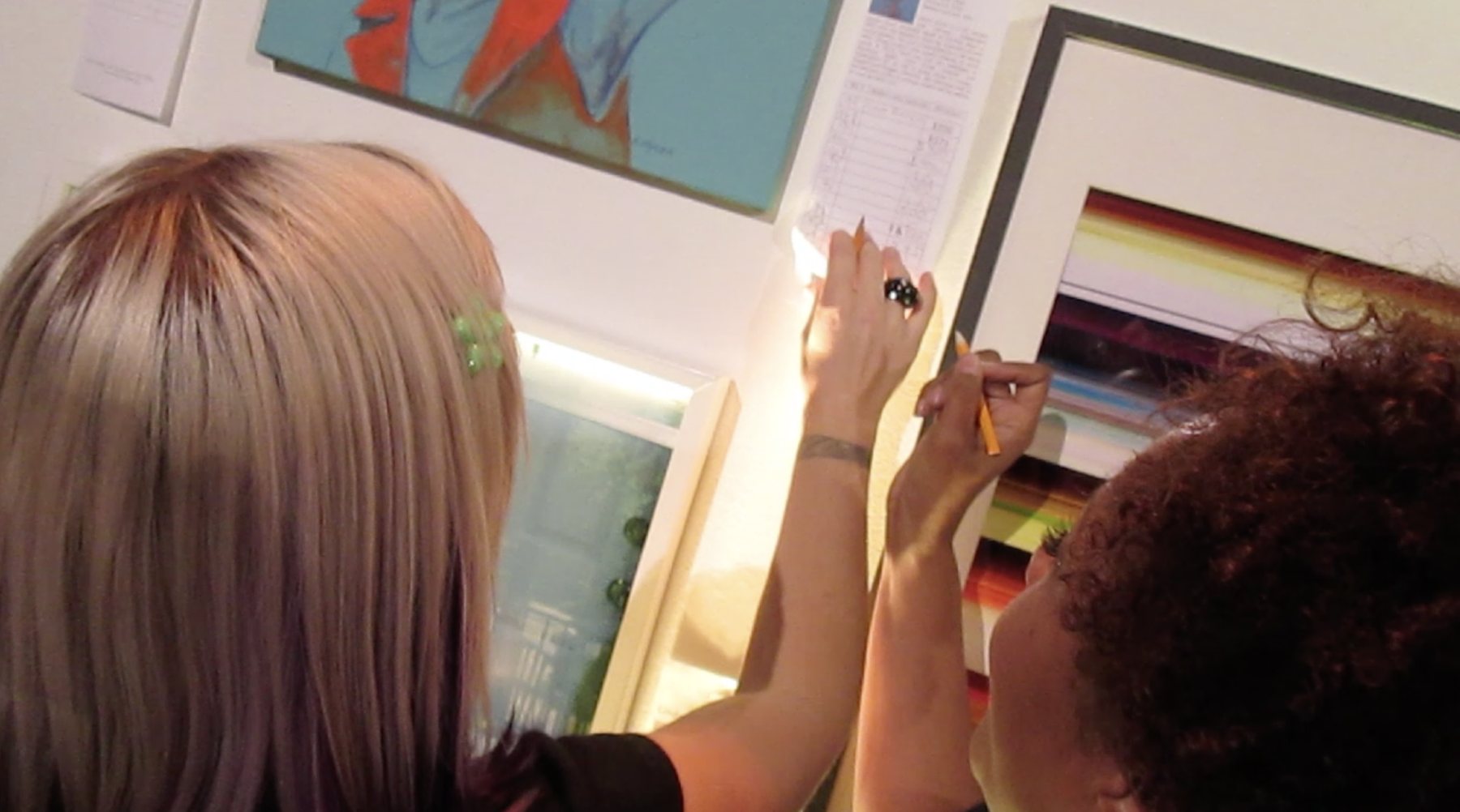 Two bidders with pencils writing bids on the bid sheet for Dana Ellyn's painting Angela at the 2014 Transformer auction